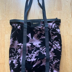 Tote från DAY ET - Snygg tote-väska från DAY ET i lila och med svart blommigt mönster i sammet. Väskan har långa svarta axelband och dragkedja upptill. Materialet är en mix av satin och sammet som ger en lyxig känsla.