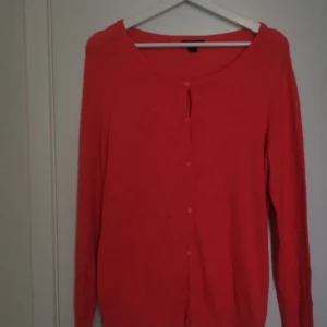 Korallröd kofta med knappar - Säljer en tunn och mjuk korallröd kofta med långa ärmar och rund halsringning. Koftan har knäppning framtill med små knappar och är perfekt att slänga över en topp. Enkel och stilren design.