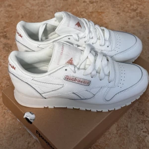 Vita Reebok Classic sneakers - Fräscha vita Reebok Classic sneakers med diskreta rosa detaljer på loggan. Helt oanvända. Skorna har snörning, rund tå och platt sula. Perfekta för dig som gillar en stilren och tidlös look. Kontakta vid frågor 🤍