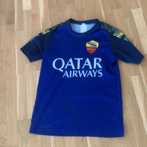 Blå AS Roma fotbollströja med Qatar Airways-tryck - Säljer en blå AS Roma fotbollströja med Qatar Airways-tryck på framsidan och namn/nummer 'Olsen 1' i gult på ryggen. Tröjan har korta ärmar med gula detaljer på axlarna och klubbmärke på bröstet. Det är 152 storlek 