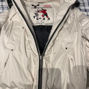 Vit dunjacka med pälskrage från Moose Knuckles - Säljer en snygg vit dunjacka från Moose Knuckles med avtagbar huva och pälskant. Jackan har flera fickor med lock och dragkedja framtill. Klassisk logga på ärmen och coolt innerfoder med hockeymotiv.