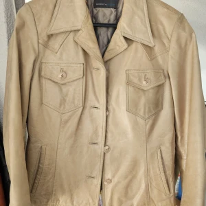 Beige skinnjacka från Rock'n Blue - Snygg beige skinnjacka med bred krage och knappar framtill. Jackan har två bröstfickor med lock och knappar samt två snedställda fickor vid sidorna. Quiltat foder på insidan för extra komfort. Etikett med storleken saknas, gissar storlek 38/40. 