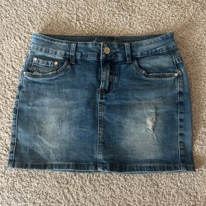 Lågmidjad jeanskjol  - Säljer en klassisk blå jeanskjol med lätt slitna detaljer och femficksmodell. Kjolen har knapp- och dragkedjestängning framtill samt bälteshällor. Perfekt för en avslappnad stil.