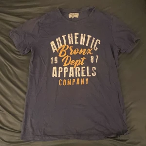 Mörkblå t-shirt från Denim Day med tryck - Mörkblå-grå t-shirt från Denim Day med stort tryck i vitt och gult framtill: 'Authentic Bronx Dept 1987 Apparels Company'. Klassisk passform och rund halsringning. Perfekt för en avslappnad stil.