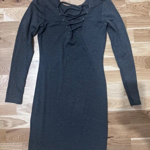Mörkgrå klänning med snörning - Säljer en mörkgrå långärmad klänning med snygg snörningsdetalj framtill vid halsen. Klänningen har en tight passform och är perfekt för dig som gillar stilrena plagg med en twist. Materialet känns mjukt och stretchigt.  Använd enstaka tillfälle och i mycket gott skick. Totallängd 85 cm  Kommer från djurfritt hem!