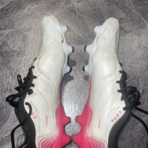 Vita och rosa Adidas Copa fotbollsskor! PRIS KAN DISKUTERAS!! - Skinnet har slitits bort som man kan se på bild 3 och 4 !!!!Säljer ett par Adidas Copa fotbollsskor i vitt med rosa detaljer och svarta skosnören. Skorna har rund tå, platt sula med dobbar och är tillverkade i syntetmaterial. Perfekta för dig som vill sticka ut på planen!