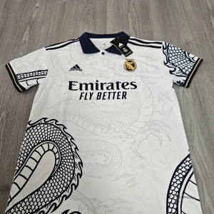 Vit Real Madrid fotbollströja med drakmönster - Säljer en vit Real Madrid fotbollströja från Adidas med mörkblå detaljer och ett stort drakmönster över hela tröjan. Tröjan har klubbmärke på bröstet, sponsortryck och klassisk krage. Perfekt för dig som vill sticka ut på planen.