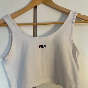 Vit croppad linnetopp från FILA - Säljer en vit croppad linnetopp från FILA med broderad logga framtill. Toppen har breda axelband och rund halsringning, perfekt för en sportig och avslappnad stil.