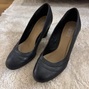 Svarta pumps med klack - Stilrena svarta pumps från River One med rund tå och bred klack. Skorna har en klassisk design i slätt material och passar perfekt till dig som vill ha en enkel men elegant sko. Klack ca 6 cm