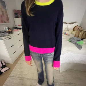 Mörkblå stickad tröja med neonrosa och neongula detaljer - Säljer en mörkblå stickad tröja med neonrosa muddar och nederkant samt neongul halsringning. Tröjan har lång ärm och en klassisk, rak passform. Perfekt för dig som gillar färgklickar i din outfit!