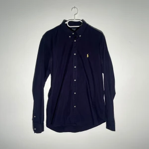Mörkblå skjorta från Polo Ralph Lauren - Klassisk mörkblå skjorta från Polo Ralph Lauren med button-down krage och gul broderad logga på bröstet. Skjortan har långa ärmar och vita knappar framtill. Perfekt för en stilren och tidlös look.
