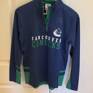 Blå Vancouver Canucks långärmad tröja NHL Signerad Av Elias Pettersson - Säljer en blå långärmad tröja från NHL med Vancouver Canucks-logga och text framtill. Tröjan har gröna detaljer vid ärmslut, nederkant och dragkedja vid halsen. Perfekt för hockeyfans som vill visa sitt lagstöd. Signerad av Elias Pettersson