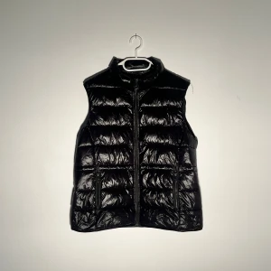 Svart glansig väst  - Säljer en svart glansig väst med hög krage och två fickor med dragkedja framtill. Västen har quiltad design och stängs med dragkedja. Perfekt att slänga över en hoodie eller tröja för extra värme.