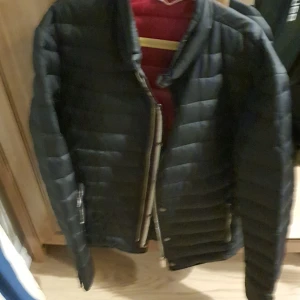 Svart pufferjacka från Aifcilanbo - Säljer en svart pufferjacka från Aifcilanbo, herrmodell i storlek M. Jackan har quiltad design, dragkedja och röd insida med serietidningsmönster. Perfekt för kyligare dagar och riktigt skön att bära.