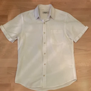 Vit kortärmad linneskjorta från Dressmann - Säljer en fräsch vit kortärmad skjorta från Dressmann i linnemix. Skjortan har klassisk krage, bröstficka och knäppning framtill. Perfekt för varma dagar och enkel att matcha med olika outfits.