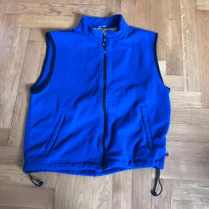 Blå fleeceväst från Tommy Hilfiger - Säljer en klarblå fleeceväst från Tommy Hilfiger med dragkedja framtill och två fickor. Västen har justerbar dragsko nedtill och svarta kantdetaljer vid ärmhål och krage. Perfekt för lager-på-lager under kyliga dagar.
