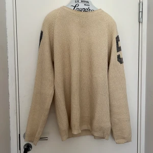 Beige stickad tröja med svarta siffror på ärmarna - Säljer en beige stickad tröja med svarta siffror på båda ärmarna. Tröjan har lång ärm och en avslappnad passform. Perfekt för dig som gillar sportig och avslappnad stil.
