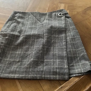 Rutig omlottkjol från H&M - Snygg rutig omlottkjol från H&M i svart, grått och vitt. Kjolen har en klassisk omlott-design med en liten metallring i midjan och är tillverkad i ett mjukt polyestermaterial. Perfekt för dig som gillar tidlösa mönster och stilren look.