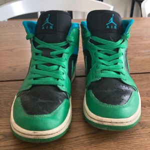 Nike Air Jordan 1 Mid i grönt och svart. Bra pris vid snabb affär.  - Säljer ett par Nike Air Jordan 1 Mid med grön och svart ovandel, blå detaljer och klassisk Air Jordan-logga på plösen. Skorna har snörning och är tillverkade i skinn med en vit sula. Perfekta för dig som gillar streetwear och sneakers med färg.