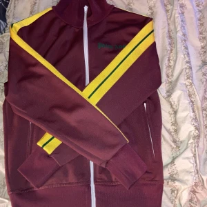 Vinröd trackjacka från Palm Angels - Säljer en vinröd trackjacka från Palm Angels med gula och gröna ränder längs axlarna. Jackan har vit dragkedja framtill, ståkrage och två fickor med dragkedja. Palm Angels-logga broderad på bröstet.