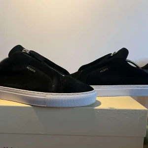 Svarta sneakers från Axel Arigato med dragkedja - Säljer ett par svarta sneakers från Axel Arigato i mocka med dragkedja på ovansidan. Skorna har vit platt sula och minimalistisk design med diskret logga på sidan. Perfekta för dig som gillar stilrena och moderna skor. Bara använda  under bröllop och bal