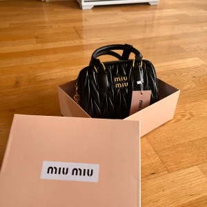 Svart handväska från Miu Miu - Säljer en svart handväska från Miu Miu med quiltad design och guldfärgade metalldetaljer. Väskan har ett lås med Miu Miu-logga och medföljande axelrem. Kommer i originalkartong och dustbag.