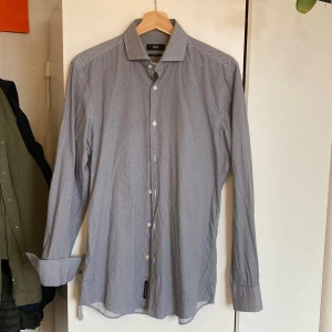 Mönstrad skjorta från BOSS - Snygg långärmad skjorta från BOSS i ett småprickigt mönster i blått och vitt. Skjortan har klassisk krage och vita knappar framtill. Liten skada (bild 3) men syns ej när skjortan är knäppt