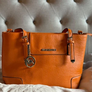 Orange handväska från Michael Kors - Säljer en snygg orange handväska från Michael Kors med klassisk MK-logga i metall. Väskan har två handtag med dekorativa spännen och är tillverkad i skinnimitation. Perfekt för dig som vill ha en färgklick till din outfit! 