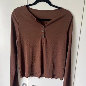 Brun ribbad långärmad topp med knappar - Säljer en brun långärmad topp med ribbad struktur och tre knappar framtill. Modellen är croppad och har en avslappnad passform. Perfekt att styla med jeans eller kjol.