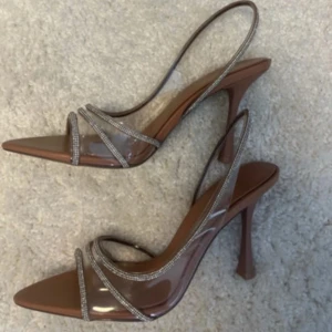 Bruna klackskor med glittriga detaljer från Zara - Säljer ett par bruna klackskor från Zara med transparenta och glittriga remmar. Skorna har spetsig tå och öppen häl, samt hög klack. Perfekta för dig som vill sticka ut med eleganta detaljer.Använda 1 gång