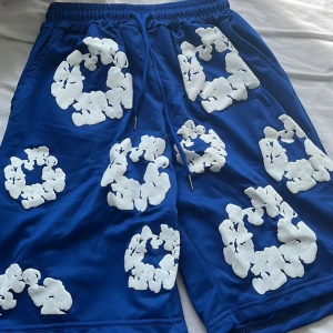 Blå shorts med vita blommiga mönster - Snygga blå shorts med unika vita blommiga mönster över hela plagget. Resår i midjan med snörning för skön passform. Perfekta för sommardagar och avslappnade tillfällen.