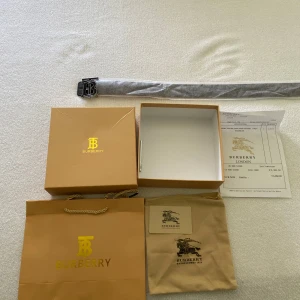 Svart bälte med silverfärgat spänne från Burberry - Säljer ett svart bälte från Burberry med klassiskt rutig mönster och stort silverfärgat 'TB'-spänne. Bältet har även Burberrys ryttare ingraverad på lädret. Kommer med originalförpackning, dustbag och påse.