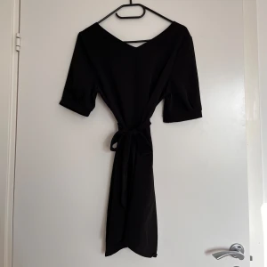 Svart tunika med knytband i midjan - En svart tunika med v-ringning och knytband i midjan. Klänningen har korta ärmar och en rak, enkel siluett som ger en stilren look. Perfekt för dig som gillar minimalistisk stil och vill ha något som funkar till många tillfällen.