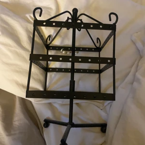 Svart smyckeshållare i metall - Snygg svart smyckeshållare i metall med flera rader och krokar för att hänga örhängen, armband eller halsband. Står stadigt på tre ben och har dekorativa detaljer upptill. Perfekt för att organisera dina smycken på ett stilrent sätt.