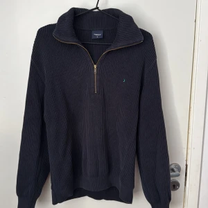 Mörkblå stickad tröja med dragkedja från Boomerang - Säljer en mörkblå stickad tröja från Boomerang i storlek M. Tröjan har halv dragkedja, hög krage och diskret broderad logga på bröstet. Ribbstickad struktur ger en snygg och klassisk look.