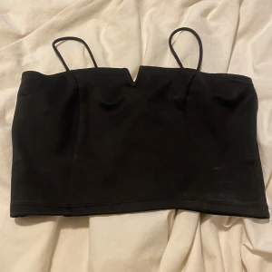 Svart croppad linnetopp från Bik Bok - Säljer en svart croppad linnetopp från Bik Bok med tunna axelband och en liten v-ringad detalj framtill. Perfekt att matcha med höga jeans eller kjol.