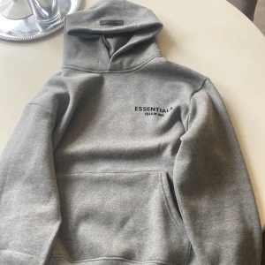 Grå hoodie från Essentials Fear of God - Säljer en grå hoodie från Essentials Fear of God med svart logga på bröstet och stor text på ryggen. Tröjan har huva och magficka, perfekt för en avslappnad stil.