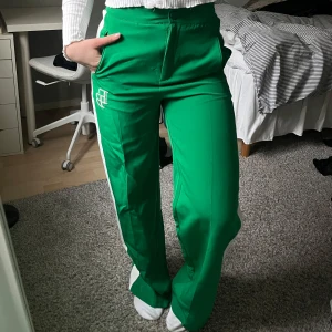 Gröna byxor med vit detalj längst sidorna - Gröna byxor i kostym-material, med raka ben och vit detalj på sidan. Byxorna har hög midja, dragkedja och knapp framtill. ”Töjbar” midja!💚