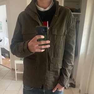 Grön fieldjacka med fickor - Perfekt för sommaren och våren. Säljer en olivgrön jacka med fyra stora fickor framtill och dragkedja. Jackan har hög krage och lång ärm, perfekt för dig som gillar en stilren och enkel look och är pe. Passar bra till jeans och sneakers 