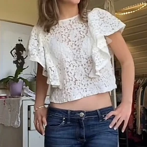 Vit  blus från Gina Tricot - Säljer en vit croppad blus från Gina Tricot i spets med blommigt mönster. Blusen har korta volangärmar och rund halsringning. 