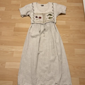 Beige långklänning med blommigt tryck från Papike Wear - Långklänning i beige linne från Papike Wear. Klänningen har korta ärmar, knytband i midjan och ett unikt blommigt tryck framtill med blommor och blad. Perfekt för dig som gillar en naturlig och avslappnad stil.