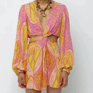 Färgglad omlottklänning med paisleymönster - Säljer en kort omlottklänning med paisleymönster i rosa, orange och gult. Klänningen har långa puffärmar, djup v-ringning och knyt i midjan. Lätt och luftig med dragkedja bak, perfekt för sommarens tillfällen. Helt oanvänd 