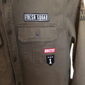 Olivgrön overshirt med patches - Säljer en olivgrön overshirt med flera coola patches, bland annat 'Fresh Squad', 'Brkln District' och stjärn- och vingeemblem. Skjortan har två bröstfickor med lock och knappar. Perfekt för dig som gillar overshirts! Den är limited edition så den går inte att få tag på längre men jag tror att den är från hm. Riktigt stilig och ovanlig overshirt! Skriv vid minsta fundering eller för fler bilder