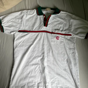 Vit piké t-shirt med gröna och röda detaljer - Säljer en vit piké t-shirt med gröna och röda ränder på krage, ärmslut och bröstet. T-shirten har korta ärmar, knappslå och en liten bröstficka med broderad logga. Tillverkad i 100% bomull. Har ett hål som man ser på bild 4.