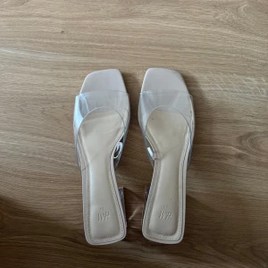 Beige klackskor med transparent rem från H&M - Säljer ett par trendiga klackskor från H&M med transparent rem och fyrkantig tå. Skorna har en beige sula och en genomskinlig klack som ger en modern look. Perfekta för dig som vill sticka ut med stilrena detaljer.