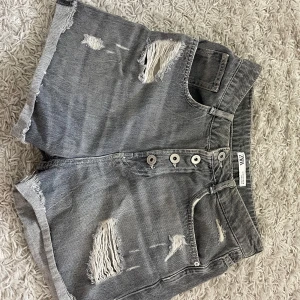 Grå jeansshorts Zara - Säljer ett par grå jeansshorts från Zara med flera slitningar och råa kanter. Shortsen har knappar framtill och klassiska fickor både fram och bak. Storlek 38