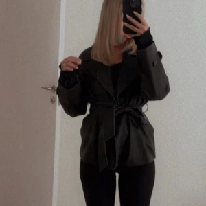 Kappa från ginatricot - Säljer en brun kappa med bälte i midjan och klassisk krage. Jackan har lång ärm och en snygg, lite oversized passform. Perfekt för dig som vill ha en stilren och trendig look.