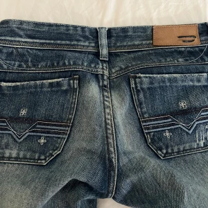Blå low waist jeans från Diesel med broderade bakfickor - Säljer ett par klassiska blå jeans från Diesel med snygga broderade detaljer på bakfickorna och en liten logotypetikett fram. Jeansen har låg midja och är lite utsvängda/raka. Enda defekten jag kan hitta är dragkedjan som är lite böjd men dem funkar jätte bra ändå.