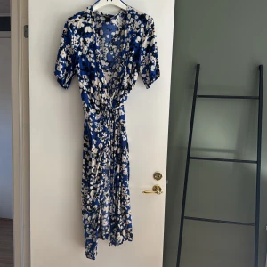 Blommig blå omlottklänning med knytband - Säljer en blå långklänning med vitt och svart blommönster. Klänningen har korta ärmar, omlottmodell och knytband i midjan för en smickrande passform. Perfekt för sommarens tillfällen!