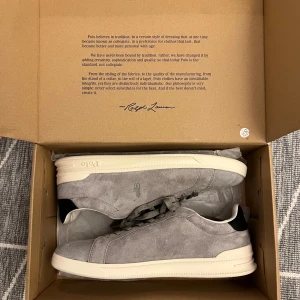 Grå sneakers från Polo Ralph Lauren - Snygga grå sneakers från Polo Ralph Lauren med vit sula och svarta detaljer vid hälen. Skorna har snörning och är tillverkade i mocka med broderad logga på sidan. Perfekta för en stilren och avslappnad look.